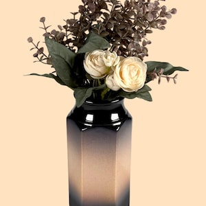 May include: A hexagonal ceramic vase with a black and tan gradient finish, containing a bouquet of artificial cream-coloured roses and dark green eucalyptus-style foliage with small brown sprigs extending from the top.
