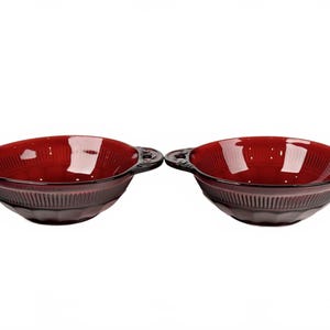 Peut inclure: Deux bols en verre rouge foncé assortis avec des poignées. Les bols présentent un motif côtelé sur la partie inférieure et un intérieur lisse et brillant. Les poignées sont petites et décoratives.