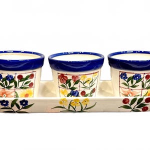 May include: Set of three ceramic flower pots with a matching tray. The pots have a white base with colorful floral designs and a blue rim. The tray is also white with floral accents, perfect for indoor or outdoor use.
