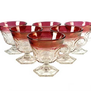 Puede incluir: Seis copas de postre de cristal transparente con borde y detalles rojo rubí. Las copas tienen base de pedestal y asa. Las copas están dispuestas en un patrón circular sobre fondo blanco.