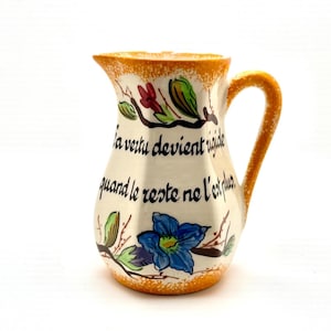 May include: Cream-colored ceramic pitcher with an orange rim and handle. The pitcher is decorated with floral designs in blue, red, and green, and features black text. The handle has an orange speckled design.