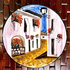 Puede incluir: Un plato decorativo redondo con una escena de calle pintada con edificios blancos, flores rojas y cielo azul. El plato tiene un borde negro y está sobre un fondo de madera tejida. La firma del artista es visible.