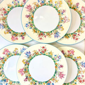 Puede incluir: Juego de seis platos con estampado floral. Cada plato presenta un fondo color crema con un vibrante diseño de corona floral en rosa, azul, amarillo y verde. Los bordes tienen un borde azul fino y el centro es blanco.