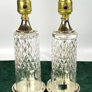 May include: Pair of vintage table lamps with clear diamond-cut glass bases, gold-toned metal accents, and brass lamp sockets. The lamps have a classic design, suitable for a living room or bedroom. Each lamp has a silver-colored base.