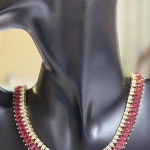 Puede incluir: Un collar y un juego de pendientes a juego en tono dorado. El collar presenta filas alternas de piedras de color rojo rubí y claras y brillantes. Los pendientes tienen un diseño similar. Las joyas se exhiben en un maniquí negro.