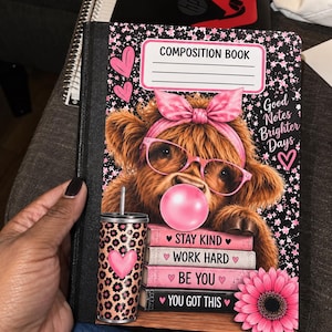 May include: A composition book featuring a fluffy brown highland cow wearing pink glasses and a pink bow, blowing a pink bubble. The cover includes a tumbler, a pink flower, and stacked books with the text: Stay Kind, Work Hard, Be You, You Got This.
