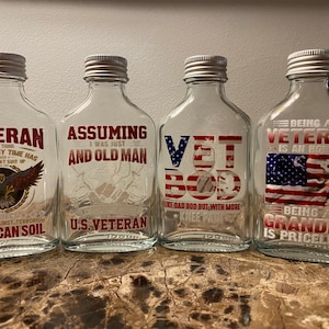 Puede incluir: Cuatro botellas de vidrio transparente con tapas plateadas. Cada botella tiene un diseño diferente con temas patrióticos rojo, blanco y azul. El texto en las botellas dice "Veteran", "Assuming I was just an old man", "Vet Bod", y "Being a Veteran is an honor, Being a Grandpa is priceless".