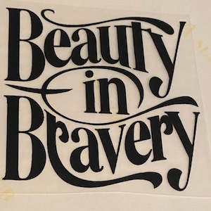 Puede incluir: Texto negro sobre fondo blanco: "Beauty in Bravery". Las palabras están en una fuente estilizada y elegante, con "in" dentro de un óvalo. El diseño es probablemente para una calcomanía o impresión.