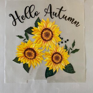 Może przedstawiać: Przezroczysta naklejka winylowa z tekstem "Hello Autumn" w czarnym stylu pisma odręcznego i trzema żółtymi słonecznikami z zielonymi liśćmi.