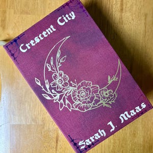 Könnte beinhalten: Ein burgunderfarbener Buchumschlag mit goldenen Lettern, auf dem "Crescent City" und "Sarah J. Maas" steht. Der Umschlag zeigt einen goldenen Halbmond mit floralen Mustern.