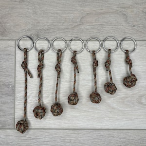 May include: A collection of seven keychains with metal rings and decorative knots. The knots are made of brown, green, and orange braided cord. The keychains are arranged on a light gray wooden surface.
