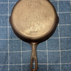 Peut inclure: Une poêle en fonte gris foncé avec une base ronde et un long manche doté d'une ouverture ovale. Le dessous est gravé avec le texte WAGNER WARE SIDNEY -O- et le numéro 1055H. La surface présente une finition métallique mate et texturée.