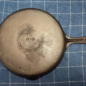 May include: A cast iron skillet with a handle. The base of the pan has the text "McCLARY", "N 9", "DRIP TOP SPIDER", and "X 617". The pan is dark grey and appears to be vintage.