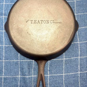 May include: A round, dark brown cast iron skillet with a long handle. The underside features the embossed text THE T. EATON CO. LIMITED and the handle is marked with the alphanumeric code X107. The surface has a textured, metallic finish.