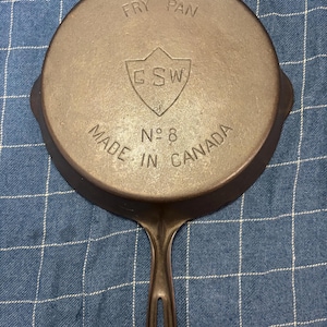 May include: A dark brown cast iron fry pan with a long handle featuring a central shield logo with the letters GSW. The text FRY PAN is embossed at the top, and the text MADE IN CANADA and N° 8 are embossed at the bottom of the pan's underside.