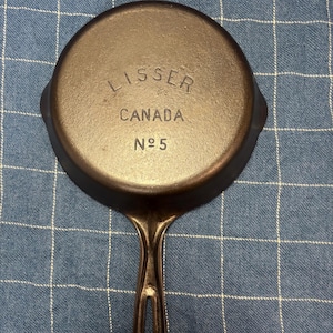 Peut inclure: Une poêle ronde en fonte avec une finition brun foncé. Le dessous présente le texte en relief LISSER, CANADA et N°5. La poêle possède une poignée unique avec une découpe ovale allongée à l'extrémité et deux petits becs verseurs sur les côtés.