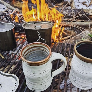 May include: Two white ceramic mugs with brown accents and a birch bark pattern sit on a metal grate over a campfire. A metal canteen with a lid is also on the grate. The mugs are filled with a dark liquid.