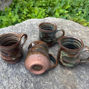 May include: Four handmade ceramic mugs with earthy tones of brown, green, and blue. Each mug has a unique shape and texture, with visible ridges and variations in color. The mugs are arranged on a gray stone surface.