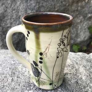 May include: A white ceramic mug with a brown rim and a black and green floral design. The mug has a handle and is sitting on a gray rock.