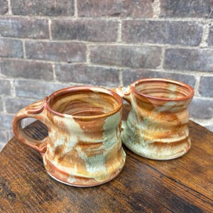 May include: Two handmade ceramic mugs with earthy tones. The mugs feature a brown, tan, and green marbled design. Each mug has a handle and sits on a wooden surface. The background is a brick wall.