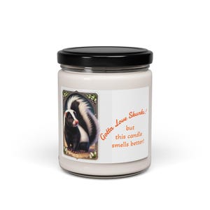 Pode incluir: Um frasco de vela branco com tampa preta. O rótulo apresenta uma ilustração de um skunk e o texto "Gotta Love Skunks! but this candle smells better!". O skunk é preto e branco com um nariz vermelho. Um item de decoração para casa.