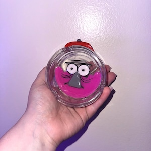 Op de afbeelding: Een heldere, ronde asbak met een cartoongezicht, roze mond, grijze neus en grote ogen. De asbak heeft een rode deksel en wordt in een hand gehouden.