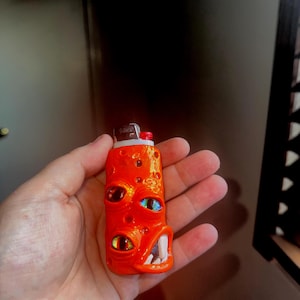 May include: An orange lighter with a monster face design. The lighter features multiple eyes in various colors and a toothy grin. The design is detailed and textured, giving it a unique, artistic appearance.