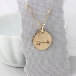 May include: A gold necklace with a round pendant featuring an arrow design. The pendant is hanging on a white background.