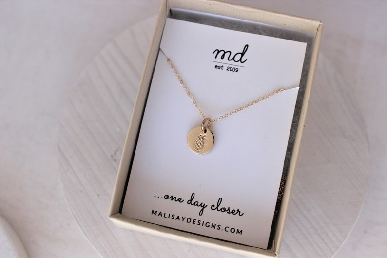 K&ouml;nnte beinhalten: Goldkette mit einem kleinen runden Anh&auml;nger mit einem Ananas-Design. Die Kette befindet sich in einer wei&szlig;en Schachtel mit dem Text "md est 2009" und "...one day closer MALISAYDESIGNS.COM".