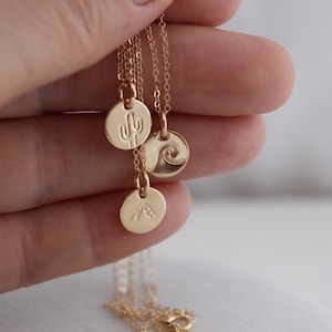 K&ouml;nnte beinhalten: Goldkette mit drei kleinen runden Anh&auml;ngern. Jeder Anh&auml;nger zeigt ein anderes Design: einen Kaktus, eine Welle und einen Berg.