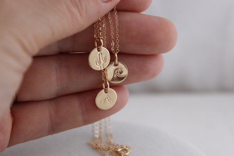 K&ouml;nnte beinhalten: Goldkette mit drei kleinen runden Anh&auml;ngern. Jeder Anh&auml;nger zeigt ein anderes Design: einen Kaktus, eine Welle und einen Berg.