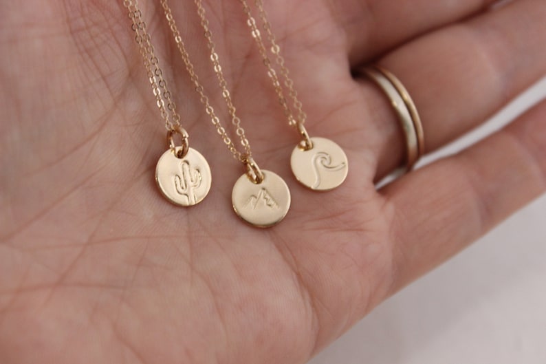 K&ouml;nnte beinhalten: Drei goldene Halsketten mit verschiedenen Anh&auml;ngern: ein Kaktus, eine Bergkette und eine Welle. Die Halsketten liegen auf einer Hand.