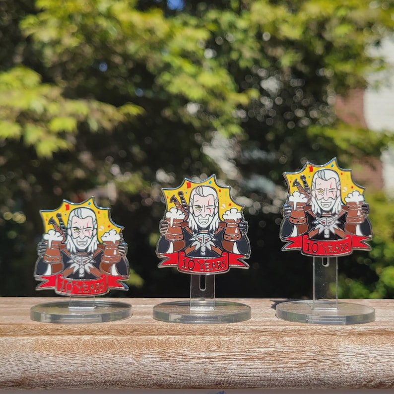 Peut inclure: Trois &eacute;pingles en m&eacute;tal argent&eacute; repr&eacute;sentant un personnage de dessin anim&eacute; tenant deux mugs de bi&egrave;re. Le personnage a les cheveux blancs et une barbe. Les &eacute;pingles sont sur des supports en plastique transparent. Les &eacute;pingles ont un fond jaune en forme d'&eacute;toile et une banni&egrave;re rouge avec le texte "10 Years".