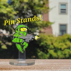 May include: A green and black enamel pin of a video game character holding a gun. The pin is on a clear acrylic stand. The text "Pin Stands!" is in the background.