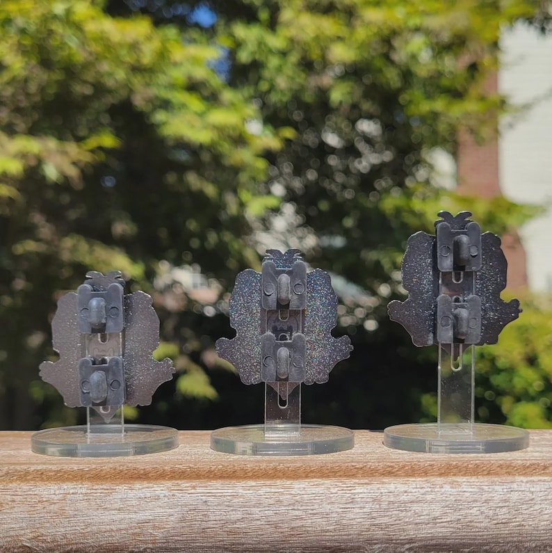 Peut inclure: Trois &eacute;pingles en &eacute;mail noir avec des accents de paillettes, chacune pr&eacute;sentant un design diff&eacute;rent, expos&eacute;es sur des supports en plastique transparent.