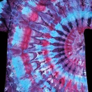 Może przedstawiać: Koszulka tie-dye w odcieniach fioletu, różu i błękitu. Wzór przedstawia spiralny wzór promieniujący ze środka, z kolorami mieszającymi się i wirującymi. Koszulka ma okrągły dekolt i krótkie rękawy.