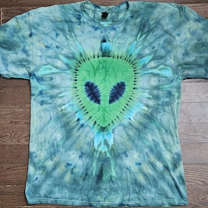 May include: A tie-dye t-shirt featuring a green alien face design with blue eyes. The shirt has a blue and green spiral tie-dye pattern. The t-shirt is a crew neck style.