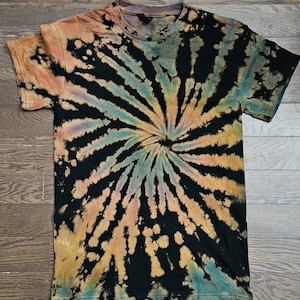 Puede incluir: Una camiseta negra teñida con un diseño de espiral. El diseño presenta una mezcla de colores como naranja, amarillo, azul y verde que irradian desde el centro. La camiseta tiene mangas cortas y cuello redondo.