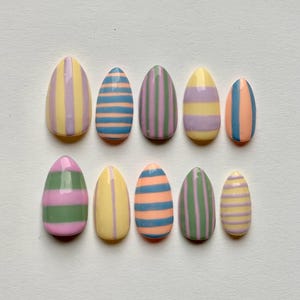 Può includere: Un set di dieci unghie finte ovali, ciascuna dipinta con una diversa combinazione di colori pastello e un design a righe. I colori includono giallo, rosa, blu, verde e viola. Le unghie sono disposte su una superficie bianca.