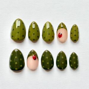 Op de afbeelding: Set van tien ovale persnagels. De meeste nagels zijn olijfgroen met donkergroene stippen. Twee nagels hebben een lichtroze basis met een rood lieveheersbeestjesontwerp. De nagels hebben een glanzende afwerking.