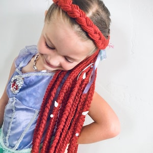May include: A young girl wearing a red braided headband and long red yarn braids with white beads. She is wearing a purple satin dress with a silver embellishment and a silver necklace.