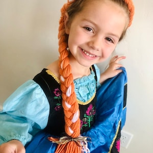 May include: A young girl wearing a blue and black dress with a white and orange yarn braid headband. The braid is long and hangs down her back. She is smiling and holding the dress up.