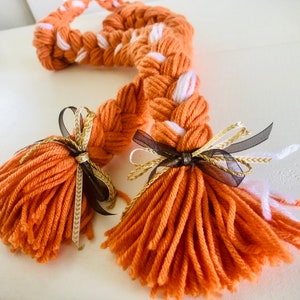 May include: Two long orange yarn braids with white accents, tied with black and gold ribbon bows. The braids are styled with fluffy orange yarn ends.