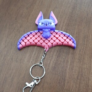 May include: A purple and pink bat-shaped keychain. The bat has a detailed design with a chain, a keyring, and a clasp. The bat's wings are pink with a scale-like pattern, and the edges are purple.