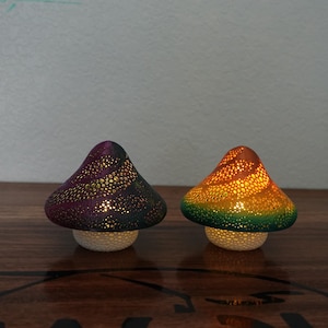 May include: Two decorative mushroom-shaped lamps. One is purple and black, the other is orange and green. Both have a perforated design on the cap, allowing light to shine through. The lamps sit on a white base.