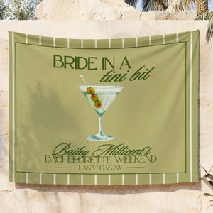 May include: A green and white banner with the text "BRIDE IN A tini bit" above a martini glass with olives. Below the martini glass is the text "Bailey Millicent's BACHELORETTE WEEKEND LAS VEGAS, NV". The banner has a green border and white stripes.