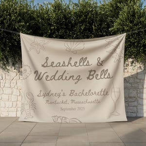 May include: A beige banner with the text "Seashells & Wedding Bells" and "Sydney's Bachelorette" with the location and date. The banner has seashell and starfish illustrations. A wine glass and lobster are also illustrated on the banner.