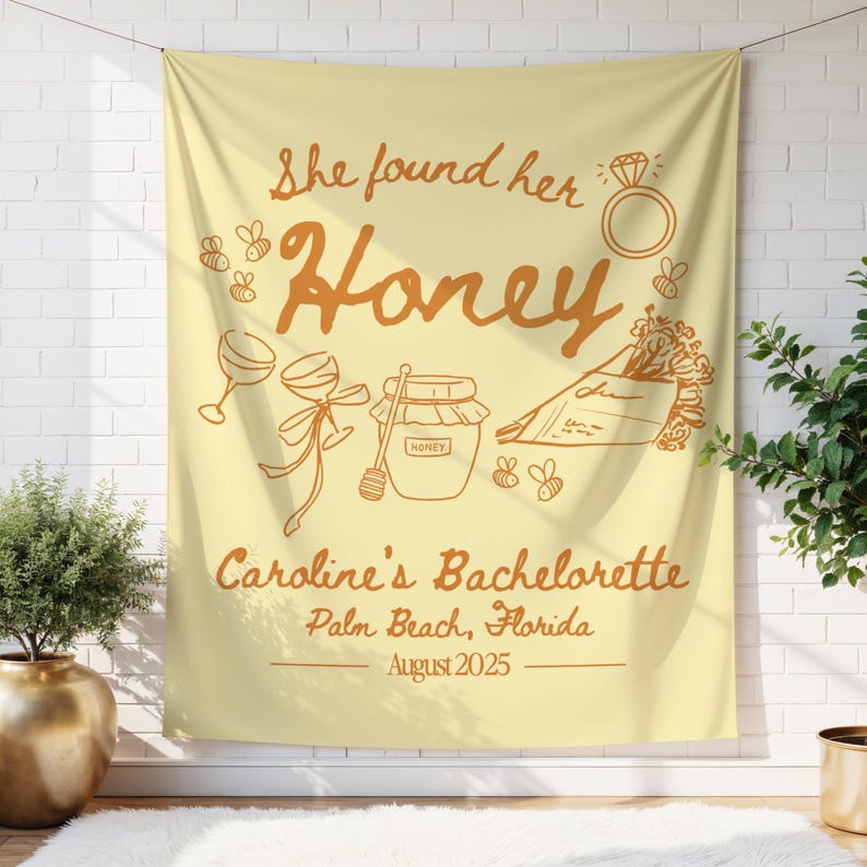 May include: A light yellow fabric wall hanging with the text "She found her Honey" in orange script. The design includes illustrations of a ring, bees, wine glasses, a jar of honey, and flowers. The text "Caroline's Bachelorette, Palm Beach, Florida, August 2025" is also included.