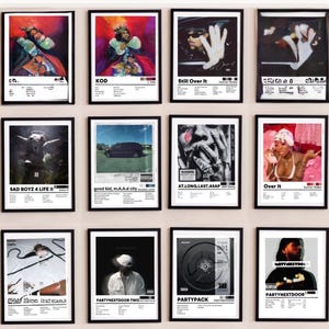 May include: A collection of twelve framed music album posters arranged in a grid. Each poster features a unique album cover art, the artist name, and a tracklist printed in black text on a white background, mimicking a minimalist design style.