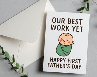 May include: A white greeting card with the text "OUR BEST WORK YET" and "HAPPY FIRST FATHER'S DAY" with an illustration of a baby wrapped in a green blanket. An envelope is next to the card.